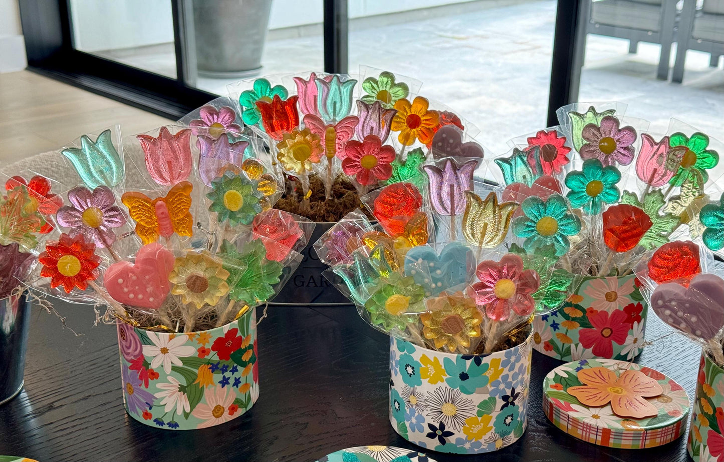 Multiple candy bouquets with handmade lollipops shaped like tulips, daisies, sunflowers, butterflies, and hearts, arranged in cylindrical floral-patterned gift boxes. The vibrant lollipops feature translucent pink, purple, yellow, green, and blue tones and are individually wrapped in clear cellophane. Displayed near a window, these cheerful edible arrangements are perfect for gifting, spring parties, or festive table decor.