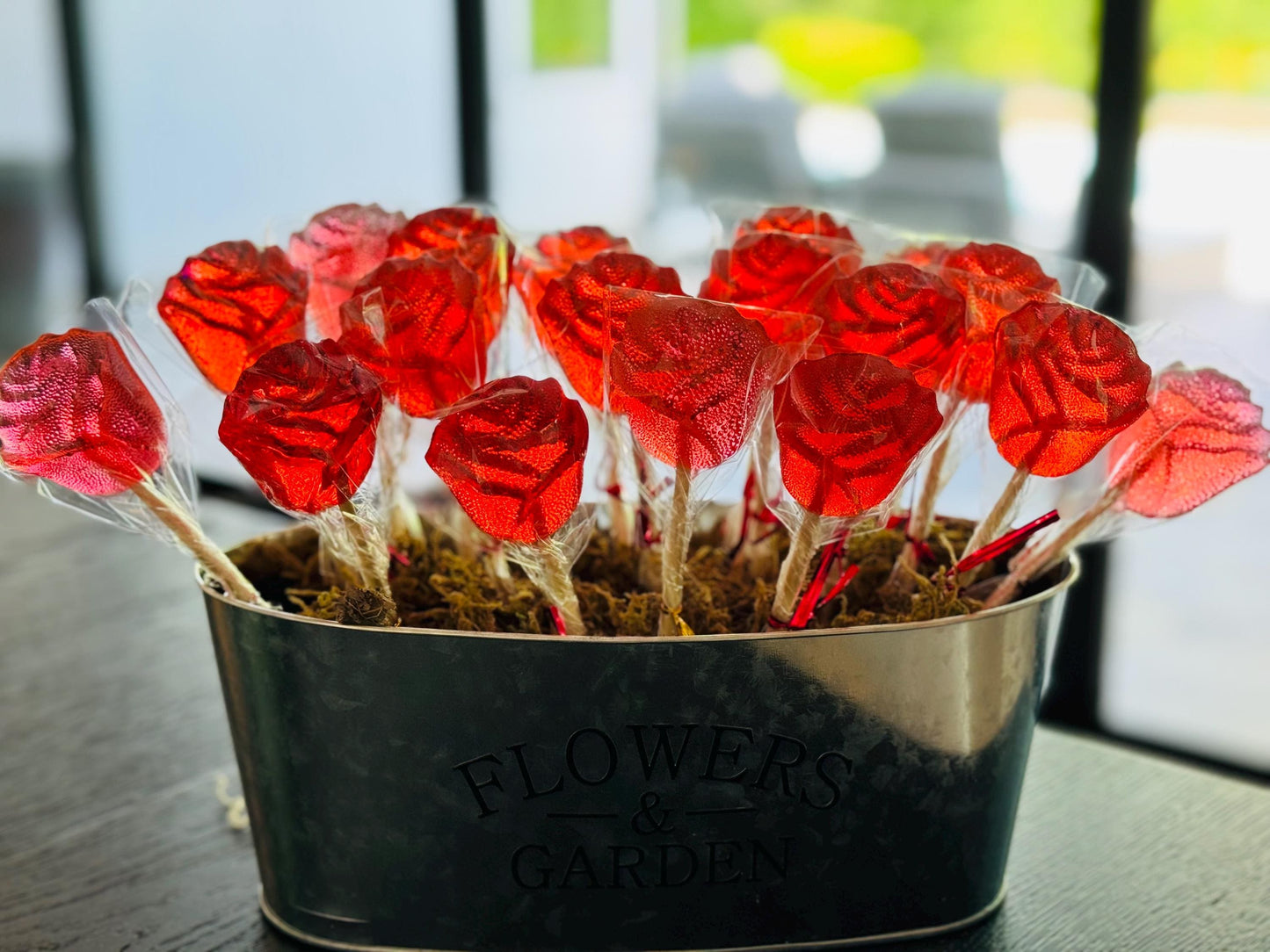 Bouquet of red rose-shaped handmade lollipops arranged in a dark metal container labeled “FLOWERS & GARDEN.” Each glossy, translucent candy is individually wrapped in clear cellophane and set upright in decorative moss to resemble a blooming rose garden. The vivid red color glows in natural light near a window. A perfect edible gift, centerpiece, or romantic party favor with vintage floral charm.
