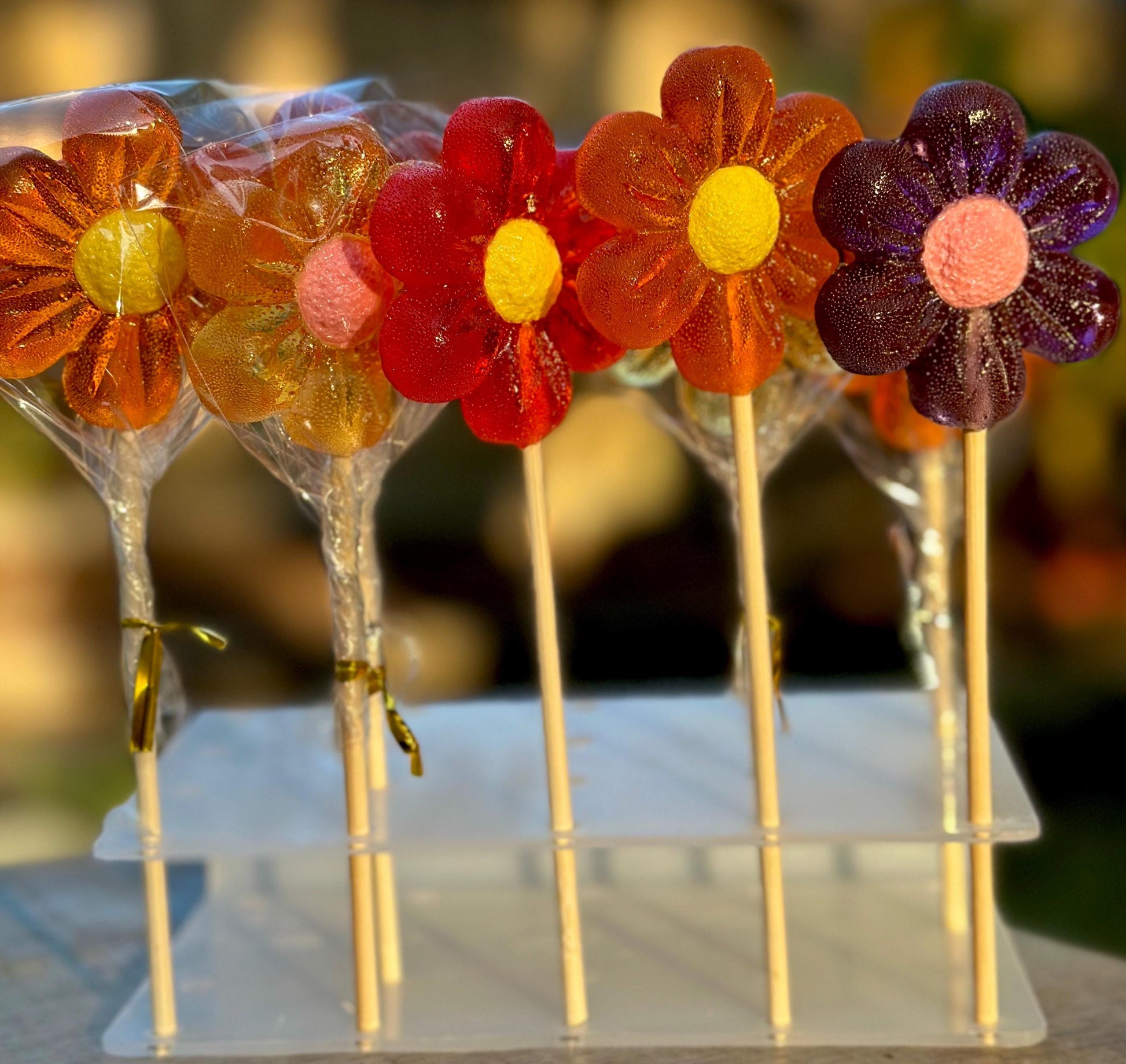A row of handmade flower-shaped lollipops in rich sunset tones—orange, red, gold, and purple—each with a bright yellow or pink center. Wrapped in clear cellophane and tied with gold twist ties, the lollipops are displayed upright on a transparent acrylic stand. Ideal for spring celebrations, baby showers, or candy bouquets.