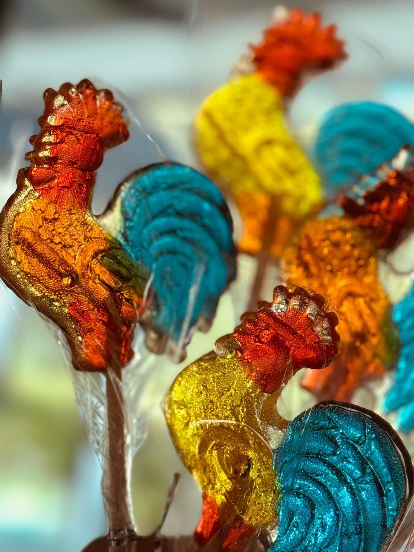 Close-up of vibrant handcrafted rooster-shaped lollipops with glossy red combs, golden yellow bodies, and teal green tails. Each lollipop is mounted on a wooden stick and wrapped in clear cellophane. The translucent candy captures light beautifully, showcasing detailed textures and nostalgic charm. Set against a softly blurred background, these artisan lollipops are perfect for gifts, party favors, or festive candy displays.