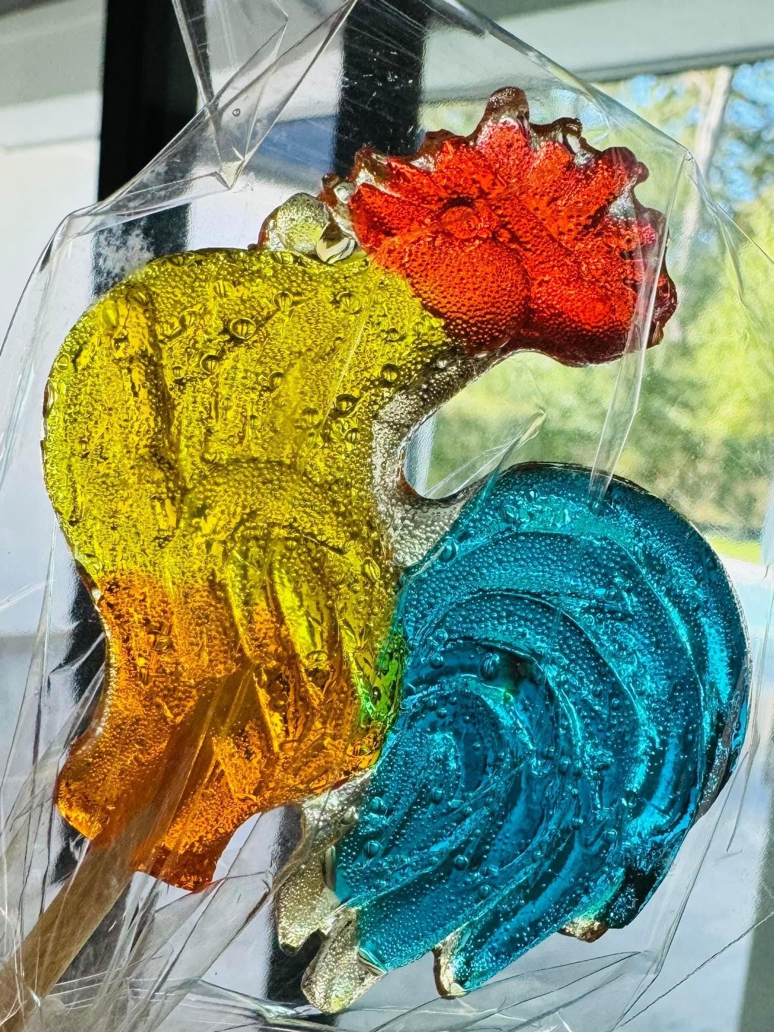 Close-up of a handcrafted rooster-shaped lollipop made from translucent hard candy with a red comb, yellow body, and vivid teal tail. Individually wrapped in clear cellophane, the candy features natural bubbling and a glossy surface that reflect its artisanal quality. Mounted on a wooden stick and shown in daylight near a window, this nostalgic rooster treat is perfect for gifting, parties, or themed candy displays.