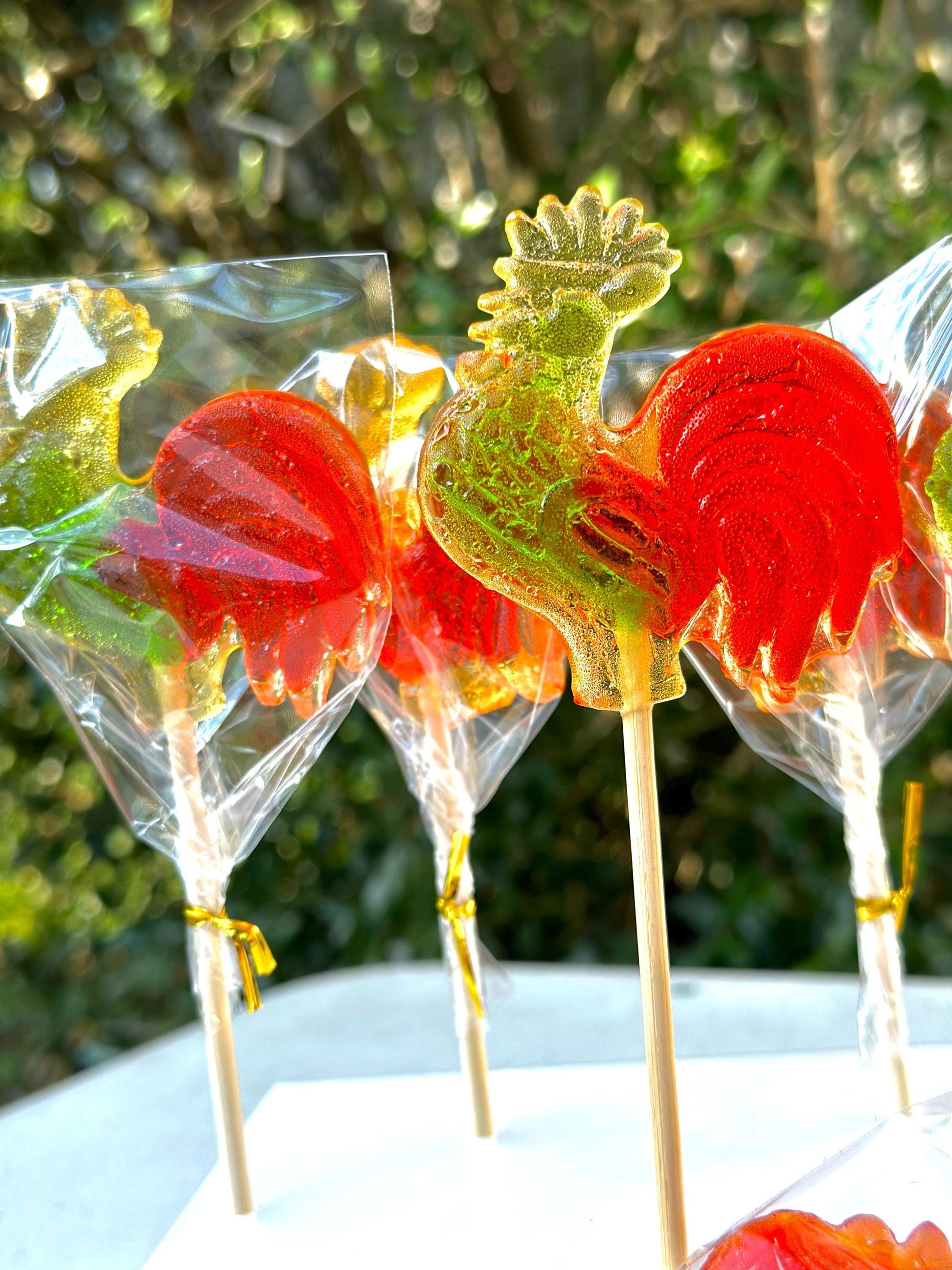 Close-up of colorful rooster-shaped lollipops displayed upright on wooden sticks. These handmade hard candies feature vibrant translucent red tails and yellow-green heads. Each lollipop is individually wrapped in clear cellophane and tied with shiny gold twist ties. Centered in the frame, the main lollipop stands out against a softly blurred green garden backdrop, creating a nostalgic, festive, and gift-ready presentation ideal for parties or holiday celebrations.