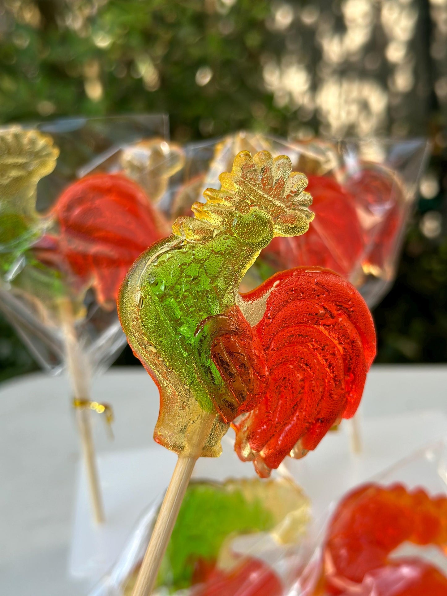 Close-up of a brightly colored rooster-shaped lollipop made from translucent hard candy, featuring a green head, golden-yellow comb, and deep red tail. Mounted on a wooden stick and individually wrapped in clear cellophane, the glossy handmade candy showcases fine texture and bubbling, emphasizing its artisanal quality. Additional lollipops and blurred green foliage in the background create a festive, nostalgic, and gift-ready presentation for parties or holidays.