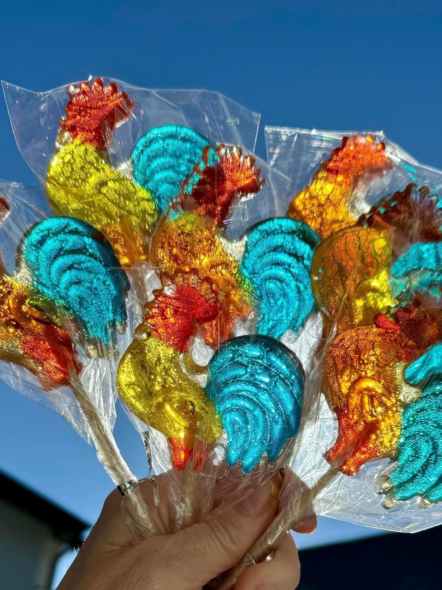 A hand holds a colorful bouquet of handmade rooster-shaped lollipops against a clear blue sky. Each hard candy features vivid translucent colors—red combs, golden yellow or orange bodies, and teal tails—and is individually wrapped in clear cellophane. The glossy finish and nostalgic rooster design make this vibrant candy arrangement ideal for party favors, gifts, or festive holiday treats.