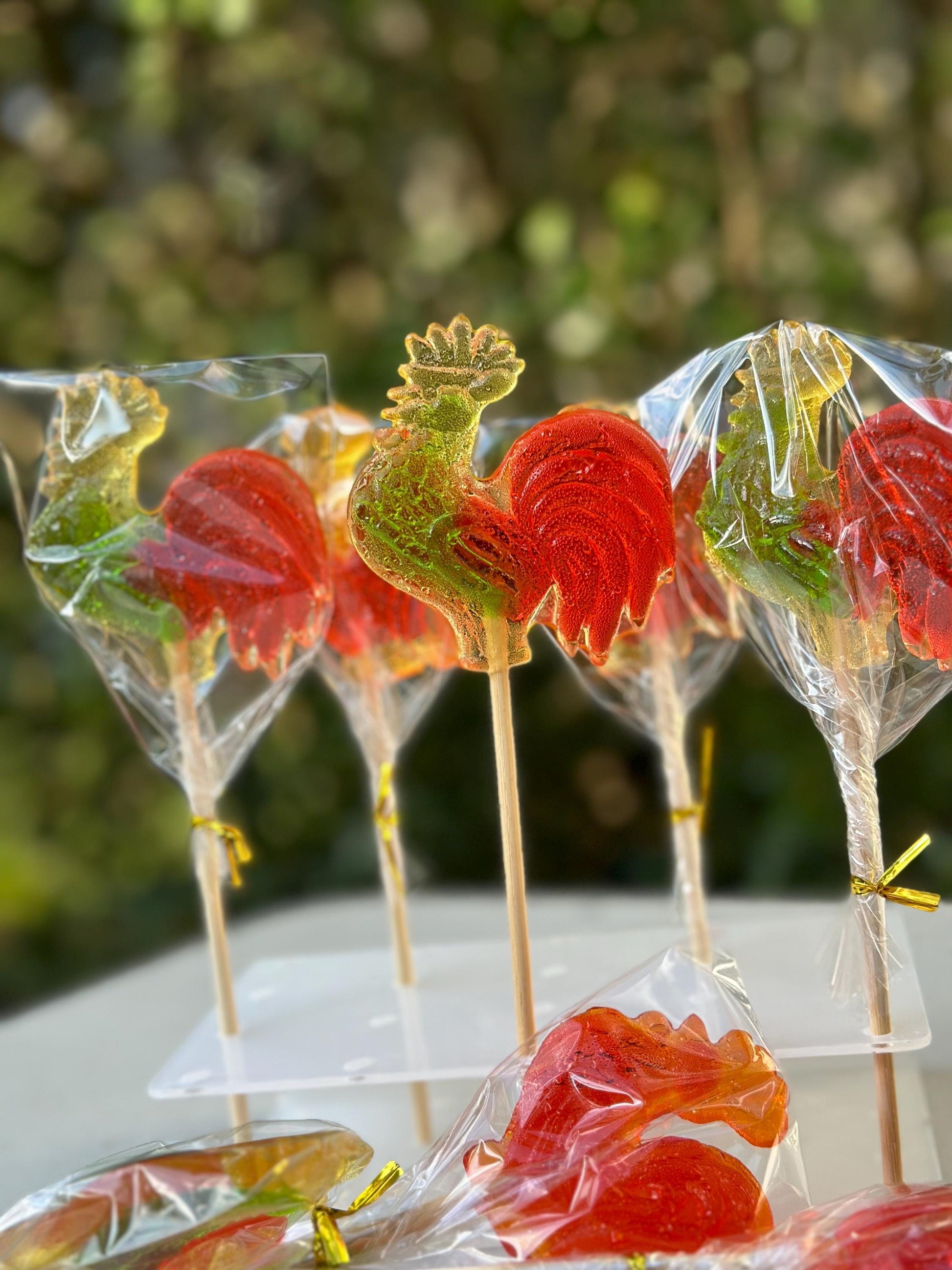 A collection of handcrafted rooster-shaped lollipops arranged upright on a transparent acrylic display stand. Each vibrant candy features a red tail and yellow-green body, sealed in clear cellophane with gold twist ties. One lollipop is placed flat in front, showcasing its detailed rooster shape and glossy surface. The softly blurred background enhances the festive, nostalgic look—ideal for parties, seasonal gifts, or holiday celebrations.