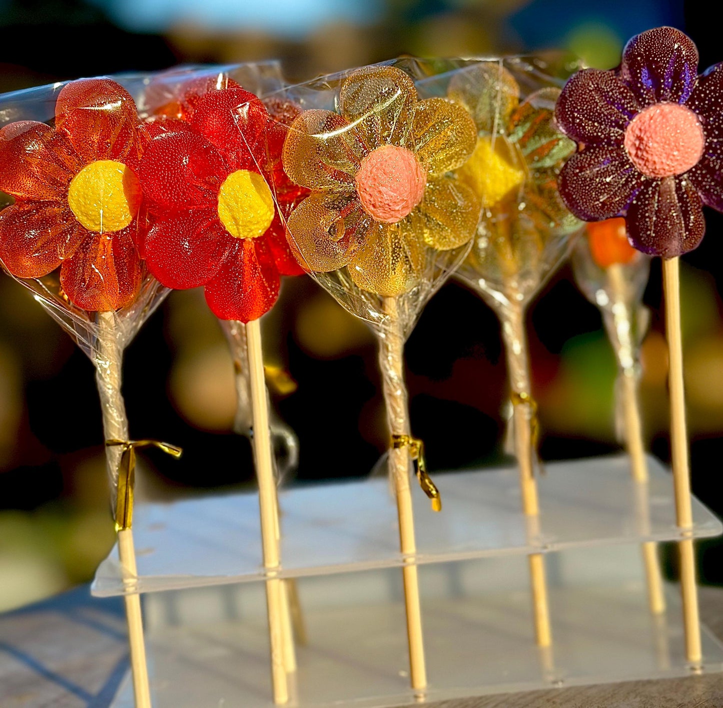 Handcrafted flower-shaped lollipops in sunset-inspired tones of red, orange, gold, and plum with vibrant yellow and pink centers. Each glossy candy is wrapped in clear cellophane and arranged on wooden sticks in a tiered display. Ideal for wedding favors, baby showers, party candy tables, or spring gifts that stand out with color and charm.