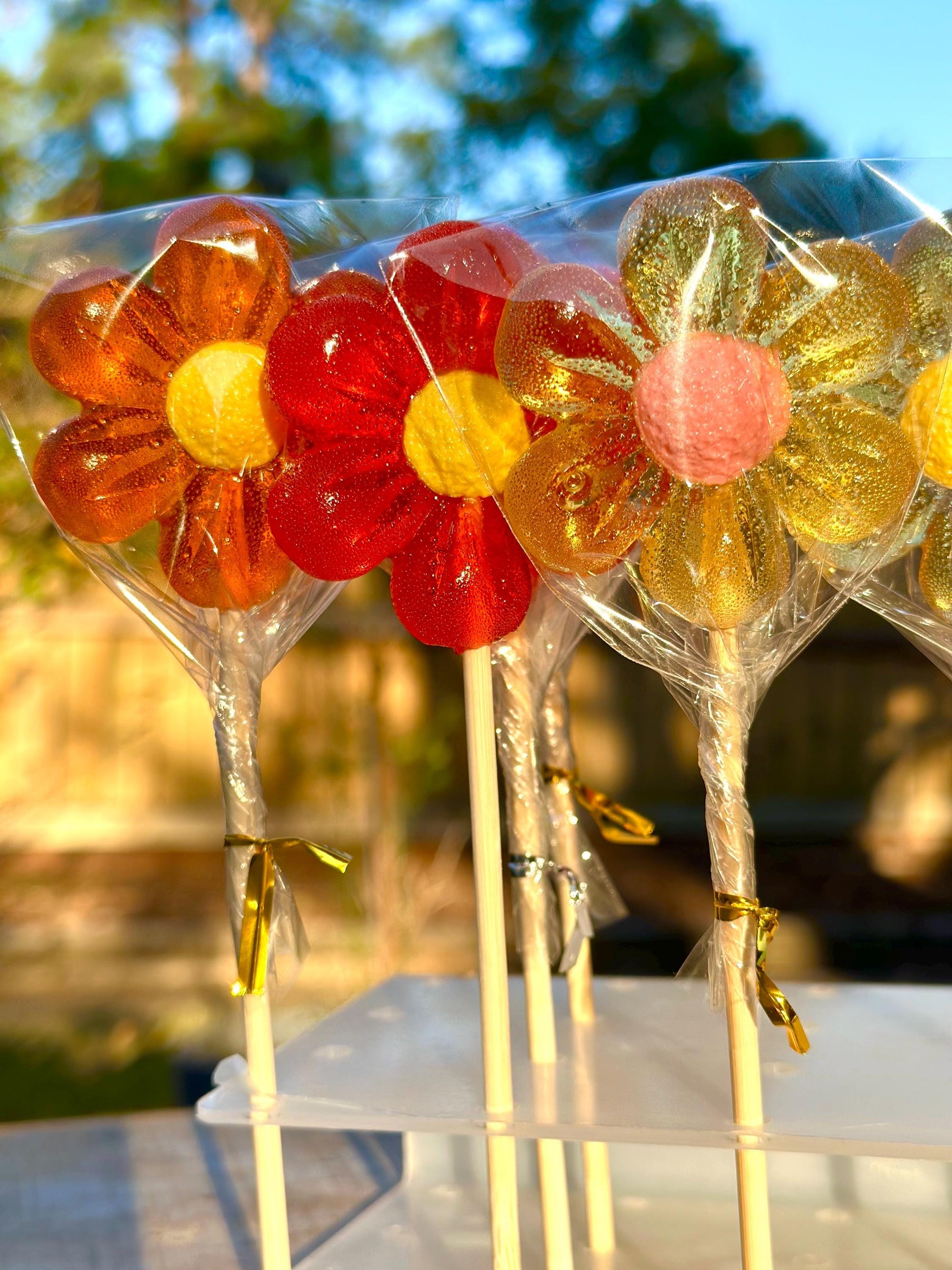 A close-up of four handmade flower-shaped lollipops with bright translucent petals in orange, red, and golden tones. Each candy has a cheerful yellow or pink center and is wrapped in clear cellophane with shiny twist ties. Displayed upright on wooden sticks, these gourmet lollipops are perfect for party favors, wedding decor, spring gifts, or Easter baskets.