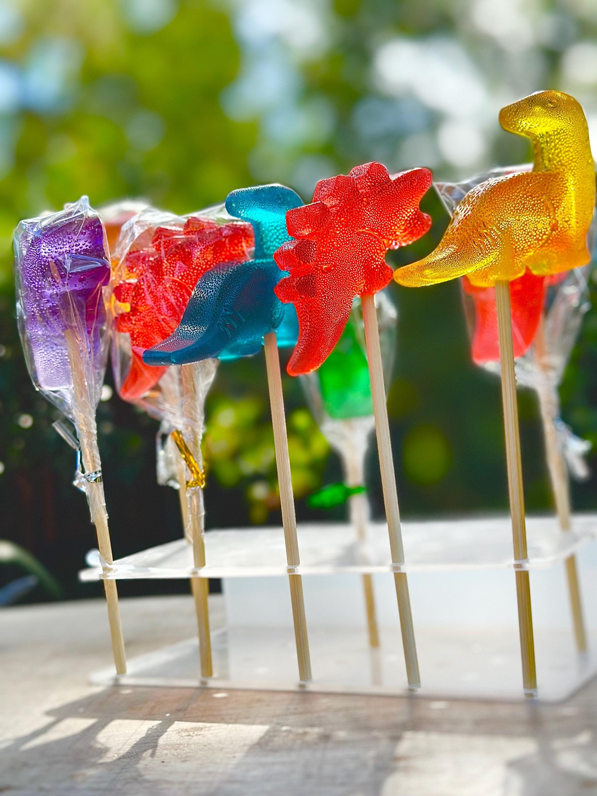 Seven bright dinosaur lollipops in vivid colors—purple, red, blue, yellow, orange—are arranged on a clear two-tier acrylic stand outdoors. Each lollipop is shaped like a different dinosaur and mounted on a wooden stick, with some wrapped in clear cellophane tied with metallic twist ties. Sunlight enhances the glossy finish, while the blurred background of trees and bokeh creates a cheerful, kid-friendly party vibe.