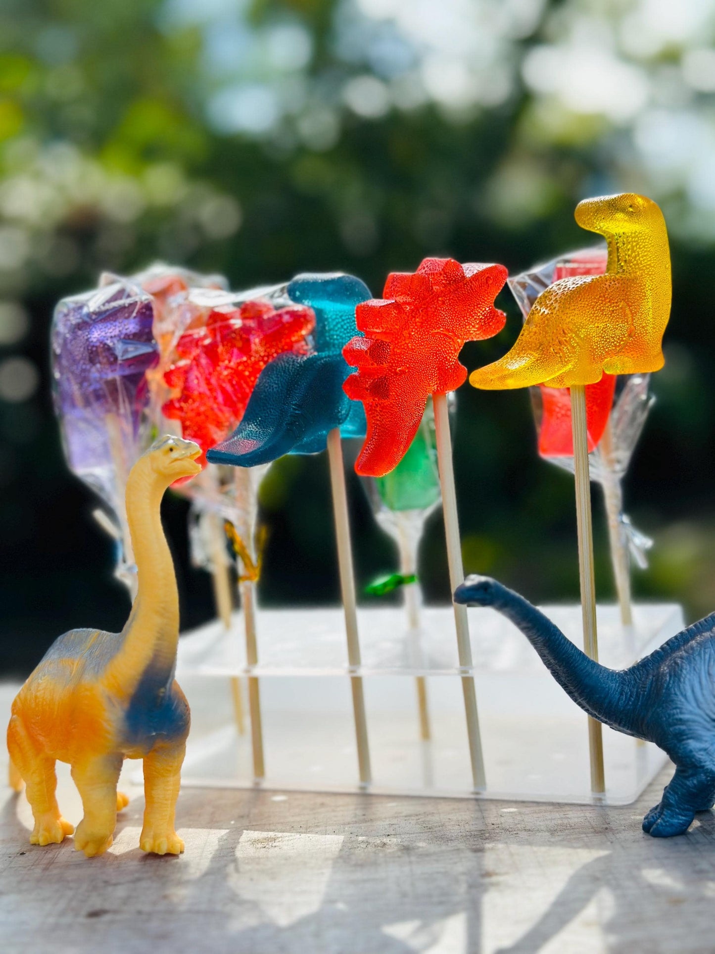 Set of six vibrant dinosaur-shaped lollipops displayed on a clear acrylic stand. Featuring detailed designs of T-Rex and Stegosaurus in red, yellow, orange, and blue, each lollipop is individually wrapped or open for display. Two toy dinosaurs in matching colors add a playful, Jurassic-themed touch. Perfect for birthday party favors, dino-themed events, or fun candy gifts for kids.