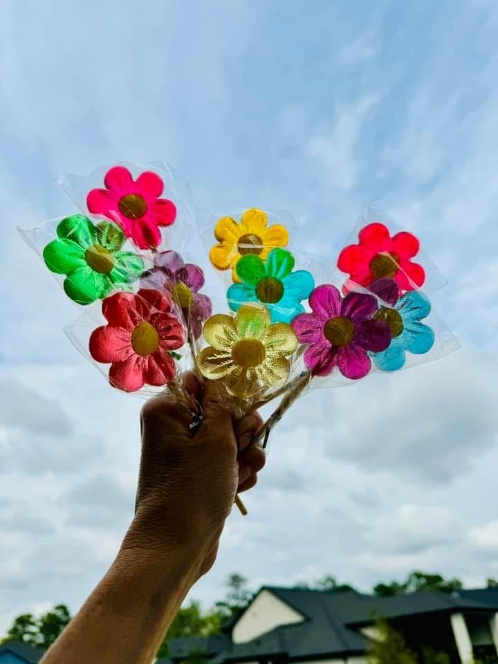 A hand holds a vibrant bouquet of handmade flower-shaped lollipops wrapped in clear cellophane against a bright sky. The lollipops resemble colorful candy flowers in pink, red, green, yellow, blue, and purple. Their translucent surface shines in natural light, creating an uplifting and cheerful effect. Ideal for candy gifting, spring celebrations, baby showers, and outdoor party decor.
