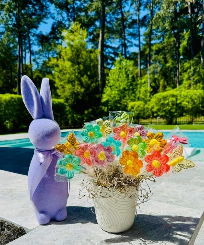 A white flowerpot holds a festive bouquet of handmade lollipops shaped like daisies and butterflies in bright translucent colors—yellow, pink, blue, orange, and purple. Each candy is individually wrapped in clear cellophane and arranged among green moss. A plush purple Easter bunny with a ribbon stands beside the bouquet near a turquoise swimming pool, creating the perfect spring or Easter candy gift display.